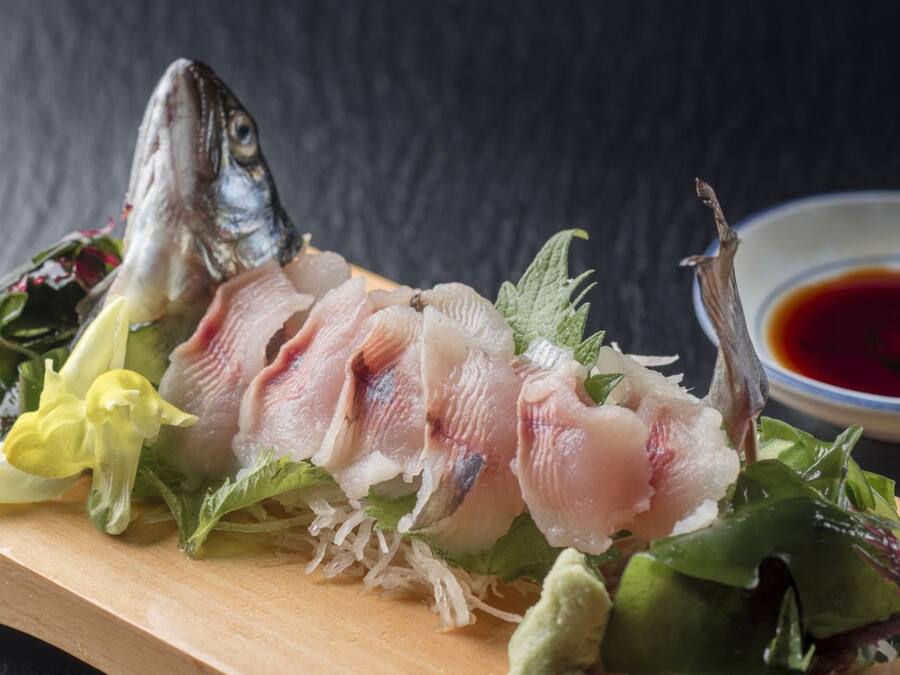 Una tabla de madera con pescado en rodajas, verduras y un recipiente pequeño con salsa.