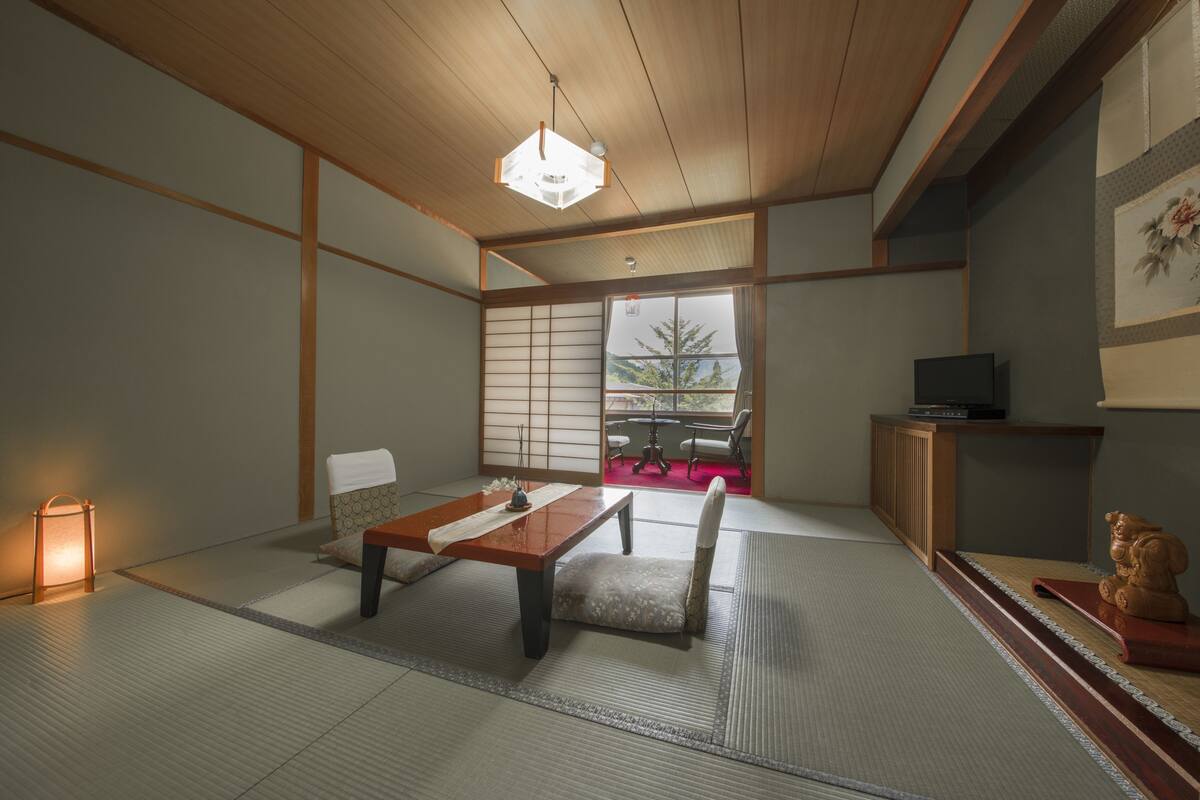 A traditional Japanese room with tatami flooring, a low table, a cushioned bench, a hanging light fixture, and a view of the outdoors through shoji screens.