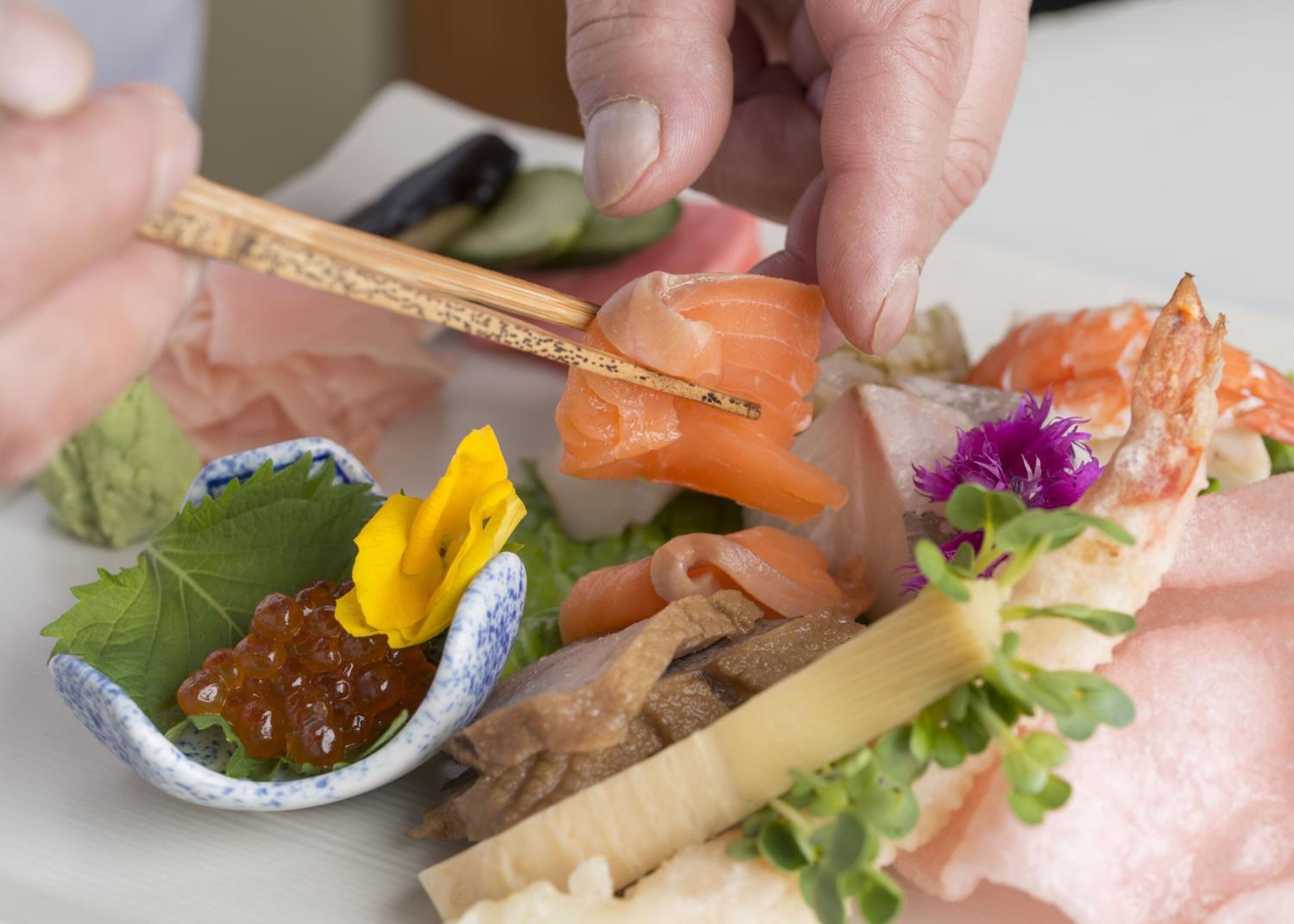 A variety of sushi and sashimi on a plate, including salmon, shrimp, and other seafood, with chopsticks and a small bowl of pickled vegetables.