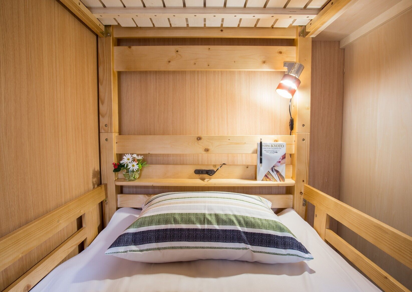 A wooden bunk bed with a bedside table, a lamp, and a book titled 'Amazing Kyoto'.