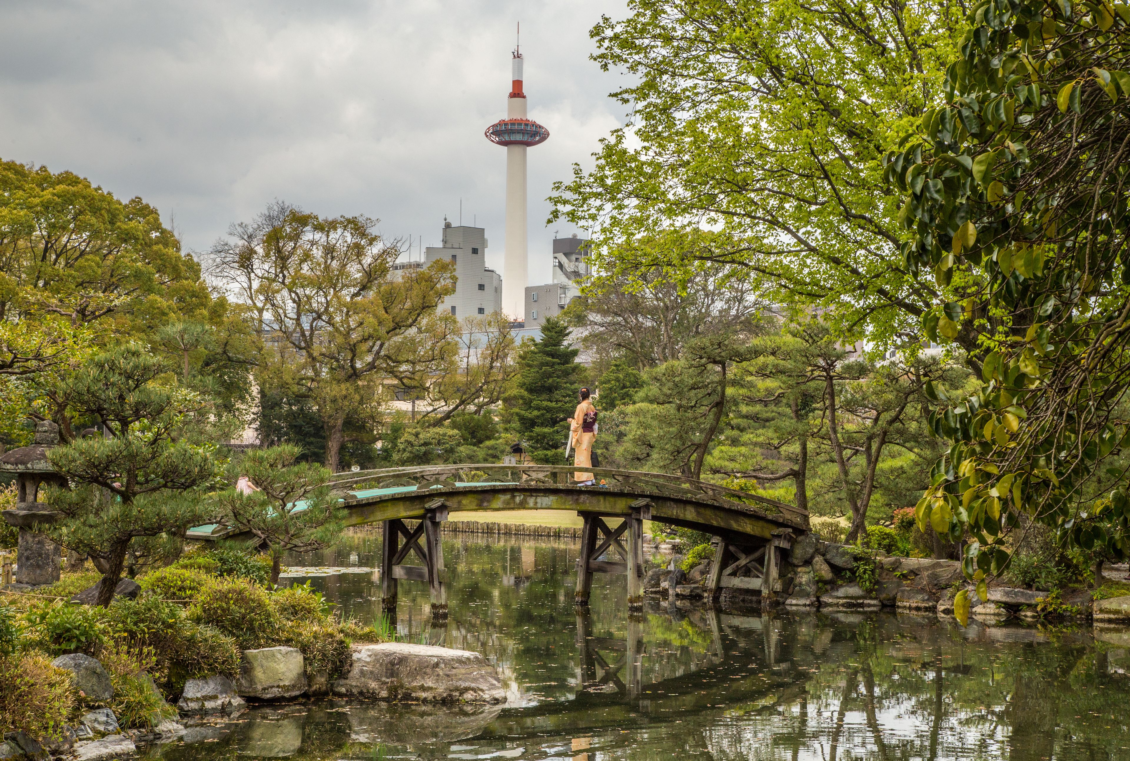 A traditional Japanese garden with a stone bridge over a pond, surrounded by lush greenery and a tower in the background.
