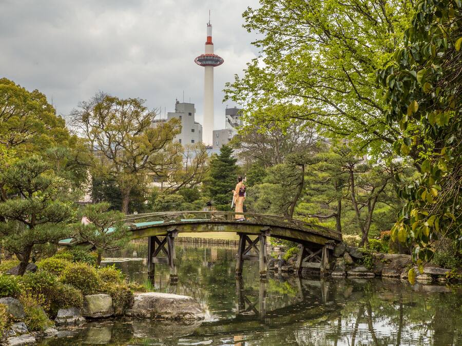 A traditional Japanese garden with a stone bridge over a pond, surrounded by lush greenery and a tower in the background.