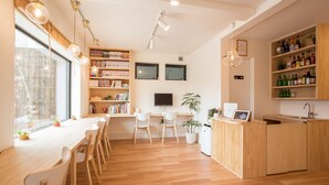 A modern, well-lit room with a wooden dining table, chairs, a kitchen area, and shelves with books and bottles. - Mulan Hostel (Kyoto)