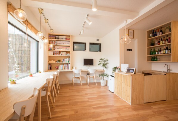 A modern, well-lit room with a wooden dining table, chairs, a kitchen area, and shelves with books and bottles. - Mulan Hostel (Kyoto)