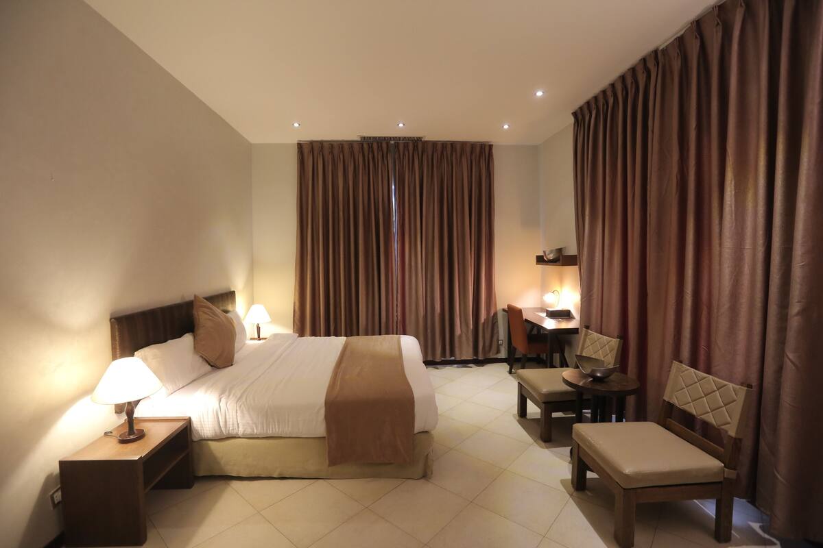 A hotel room with a bed, bedside table, lamp, chair, and a desk with a chair.
