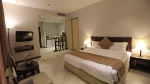A hotel room with a large bed, bedside tables with lamps, a TV mounted on the wall, a dining area with chairs, and a kitchen in the background.
