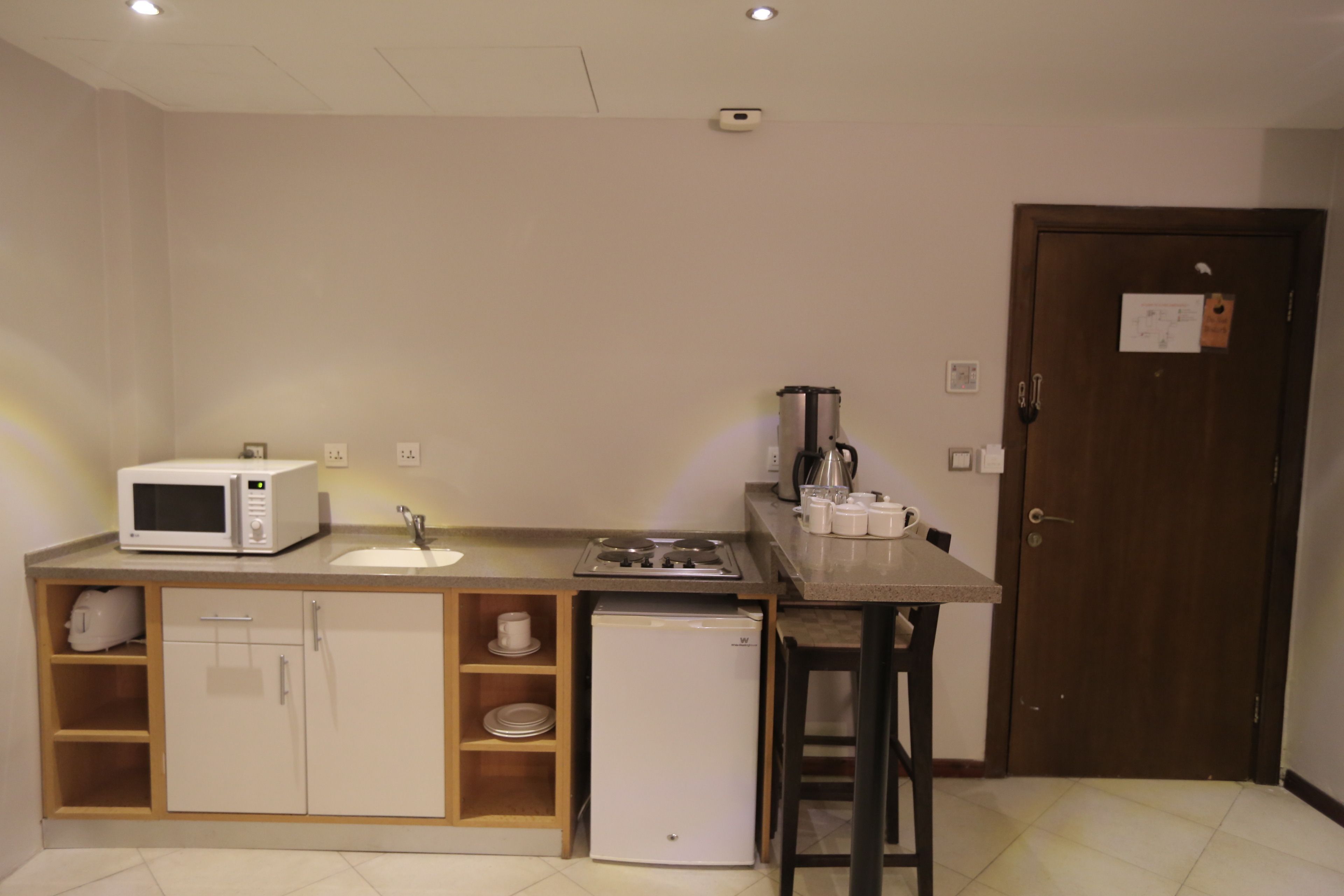 Eine kleine Küche mit Mikrowelle, Spüle und kleinem Kühlschrank. Es gibt einen Holzkasten mit Regalen und eine Arbeitsplatte mit Wasserkocher und Tassen. Im Hintergrund ist eine Tür mit einem Zettel zu sehen.