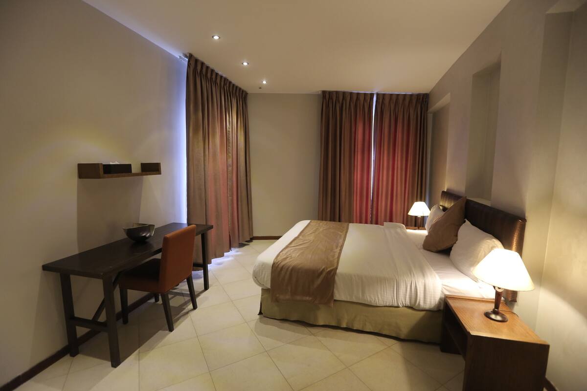 A hotel room with a bed, a desk, a chair, a lamp, and curtains.