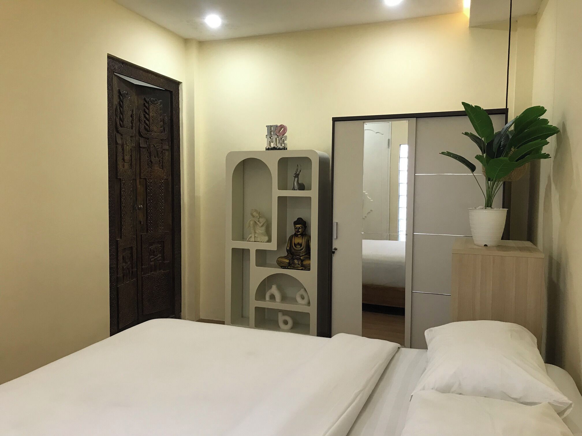 Un dormitorio con cama, armario, espejo, planta en maceta y aparador decorado con estatuas religiosas.
