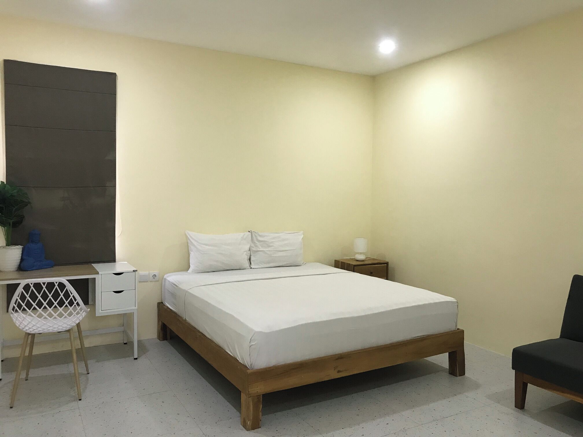 A bedroom with a wooden bed, white bedding, a desk with a chair, a small nightstand, and a potted plant.