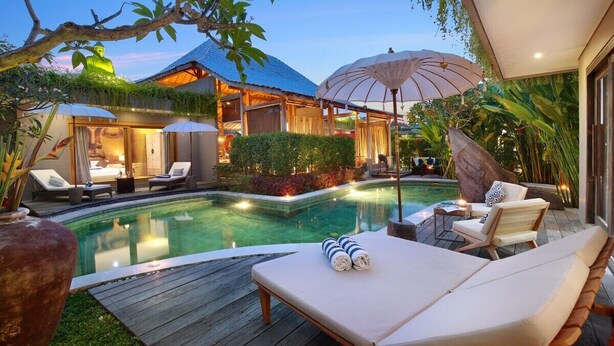 A poolside area with lounge chairs, a large umbrella, and a building with a tiled roof. - Gahana Bali Villas (Seminyak)