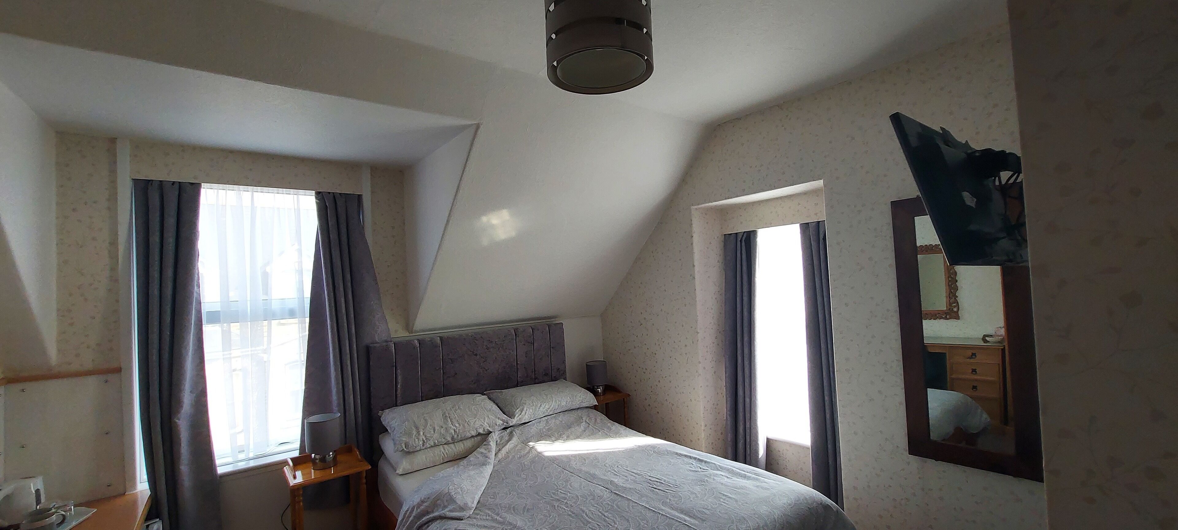 A bedroom with a bed, two bedside tables, a lamp, a mirror, and two windows with curtains.
