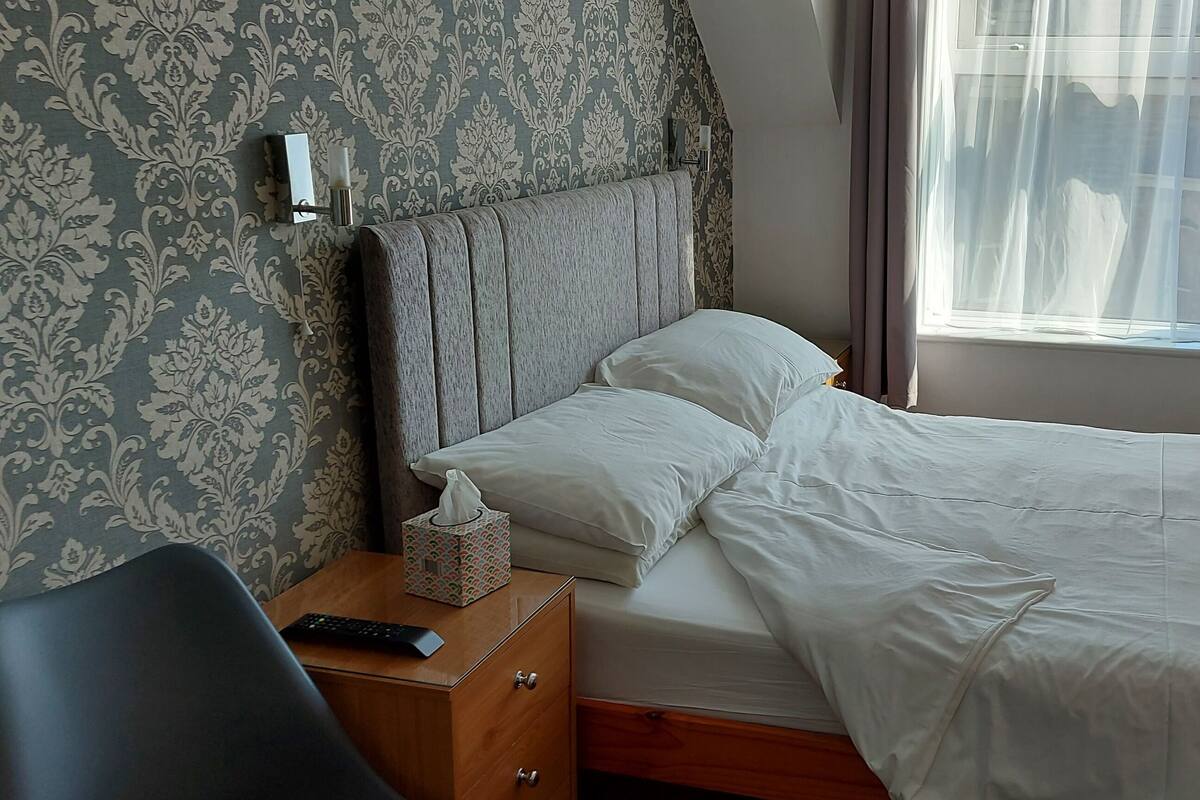 A neatly made bed with white linens, a bedside table with a remote control and a tissue box, and a patterned wallpaper on the wall.