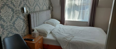 A neatly made bed with white linens, a bedside table with a remote control and a tissue box, and a patterned wallpaper on the wall.