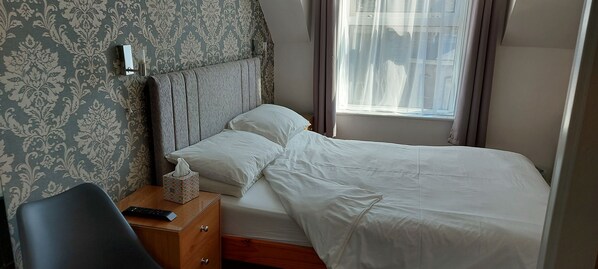 A neatly made bed with white linens, a bedside table with a remote control and a tissue box, and a patterned wallpaper on the wall. - Invernook Hotel (Newquay)