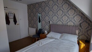 A bedroom with a bed, a chair, a nightstand, a mirror, and a door with towels hanging. - Invernook Hotel (Newquay)