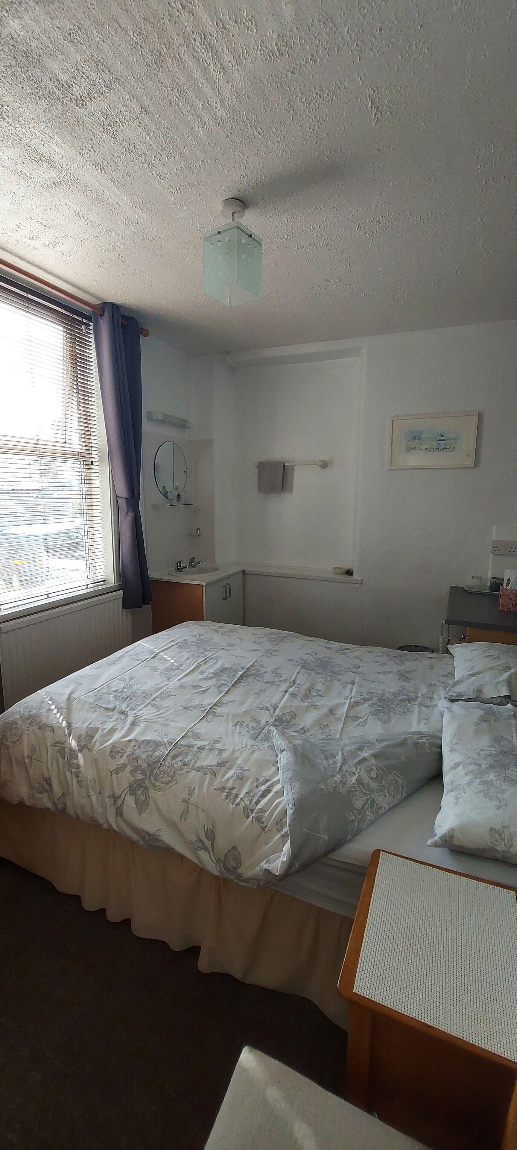 Une chambre d’hôtel avec un grand lit, une table de chevet, une petite table, une fenêtre avec des rideaux, un miroir fixé au mur, un radiateur et un luminaire au plafond.