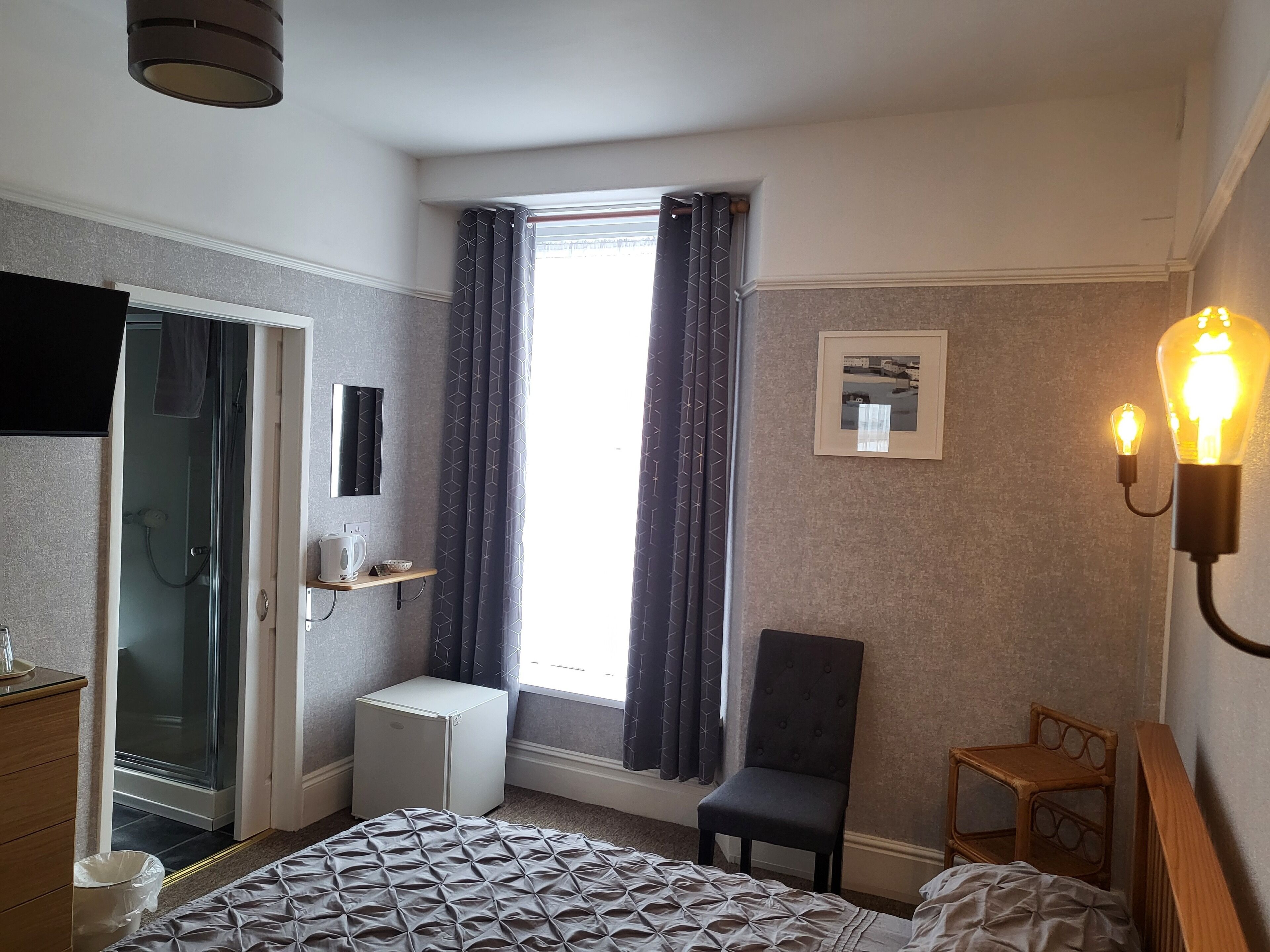 A bedroom with a bed, a chair, a small table, a heater, a window with curtains, and a picture on the wall.