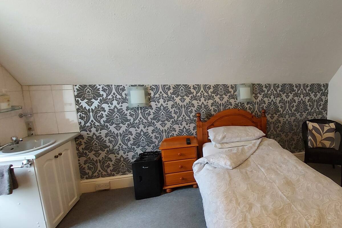 A small, neatly arranged bedroom with a single bed, a bedside table, a chair, a dresser, and a wall-mounted lamp.