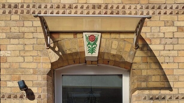 A white door with a glass panel, flanked by two potted plants, a brick wall with decorative tiles, and a sign indicating the building is an inn. - Invernook Hotel (Newquay)