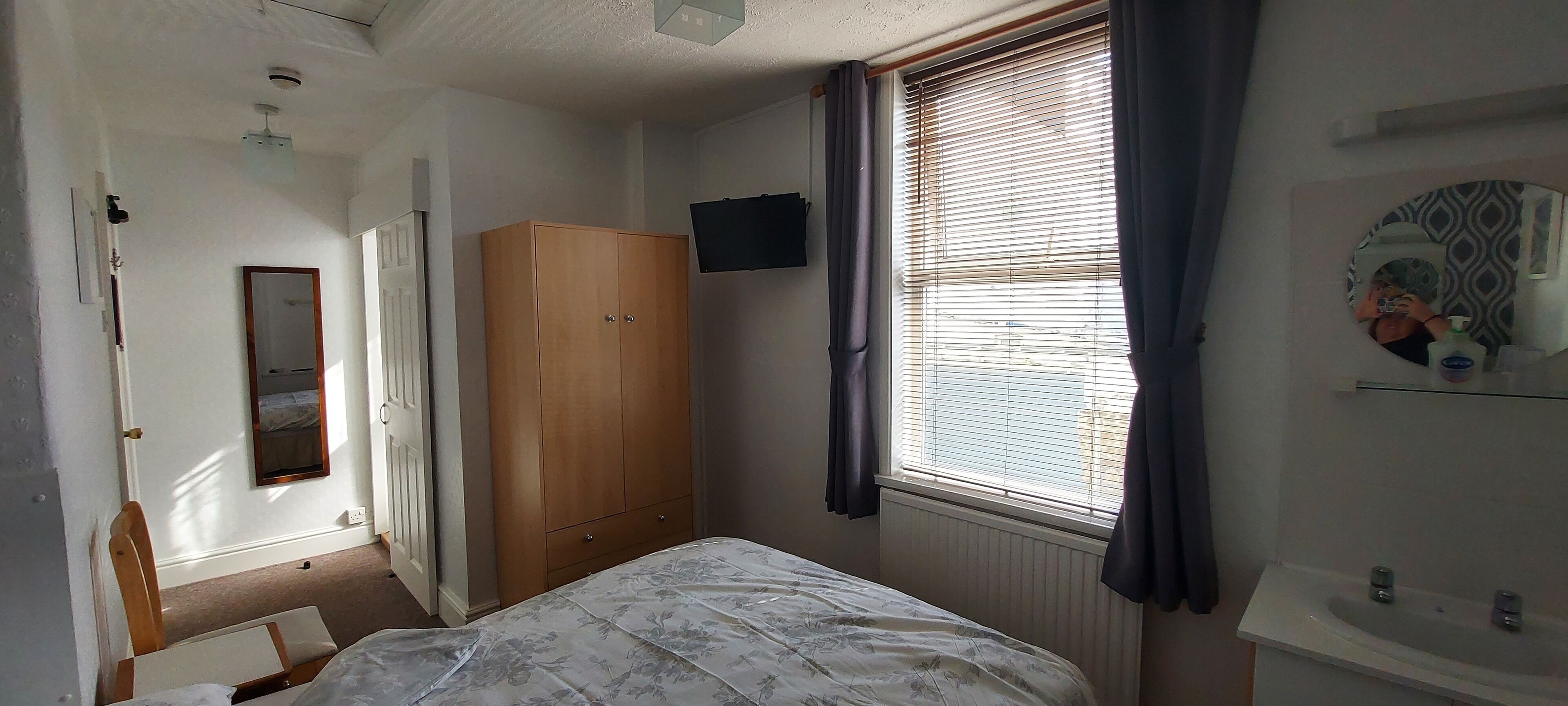 Une chambre à coucher comprenant un lit, une armoire, un miroir, une fenêtre avec des rideaux et un radiateur.
