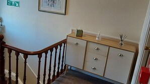 A hallway with a wooden staircase, a fire exit sign, a framed picture on the wall, a cabinet with drawers, and a fire extinguisher mounted on the wall. - Invernook Hotel (Newquay)