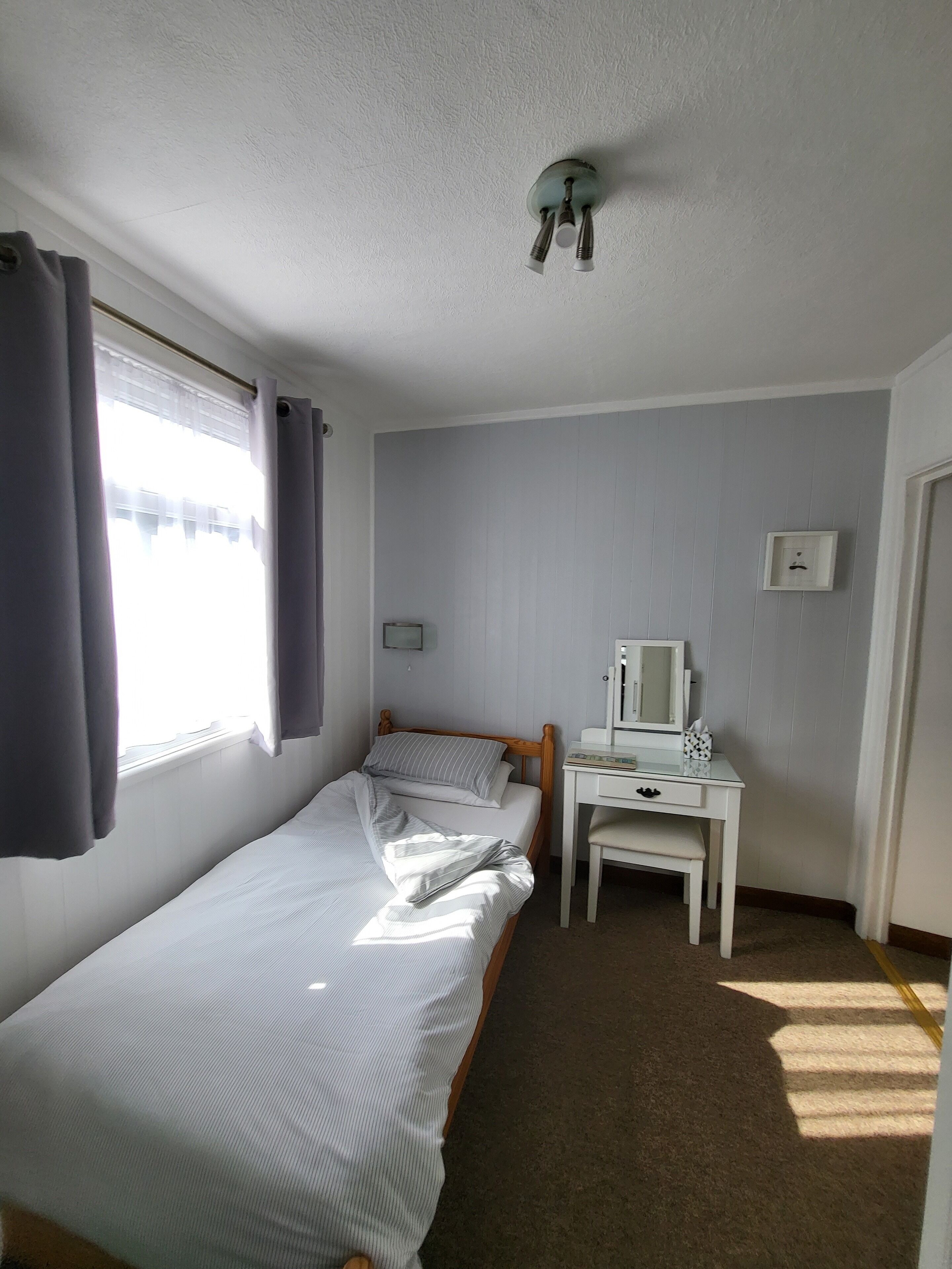 A single-bed room with a wooden headboard, a bedside table with a mirror, a small desk, and a window with curtains.