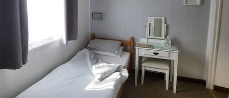 A single-bed room with a wooden headboard, a bedside table with a mirror, a small desk, and a window with curtains.