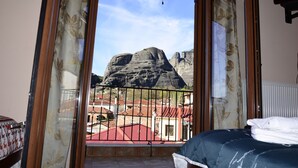 A room with a bed, a window with a view of rocky mountains, and a balcony with red-roofed buildings. - Guesthouse Lithos (Kalabaka)