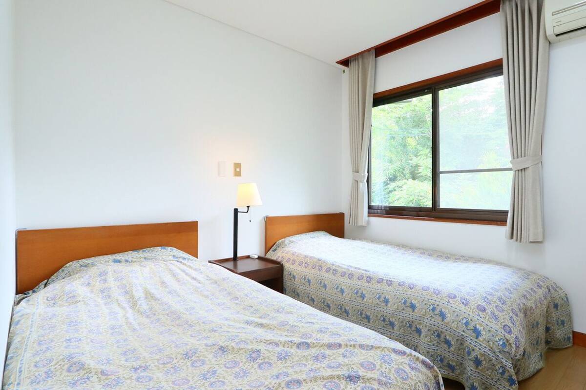 A bedroom with two beds, a nightstand with a lamp, a window with curtains, and an air conditioning unit.