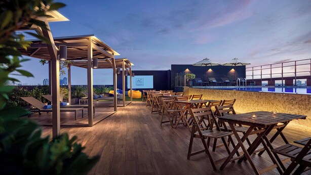 An outdoor seating area with wooden tables and chairs, a pool, and a city skyline in the background. - AC Hotel by Marriott Puchong (Puchong)
