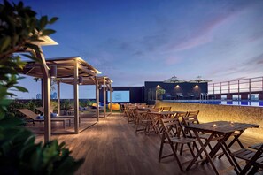 An outdoor seating area with wooden tables and chairs, a pool, and a city skyline view.