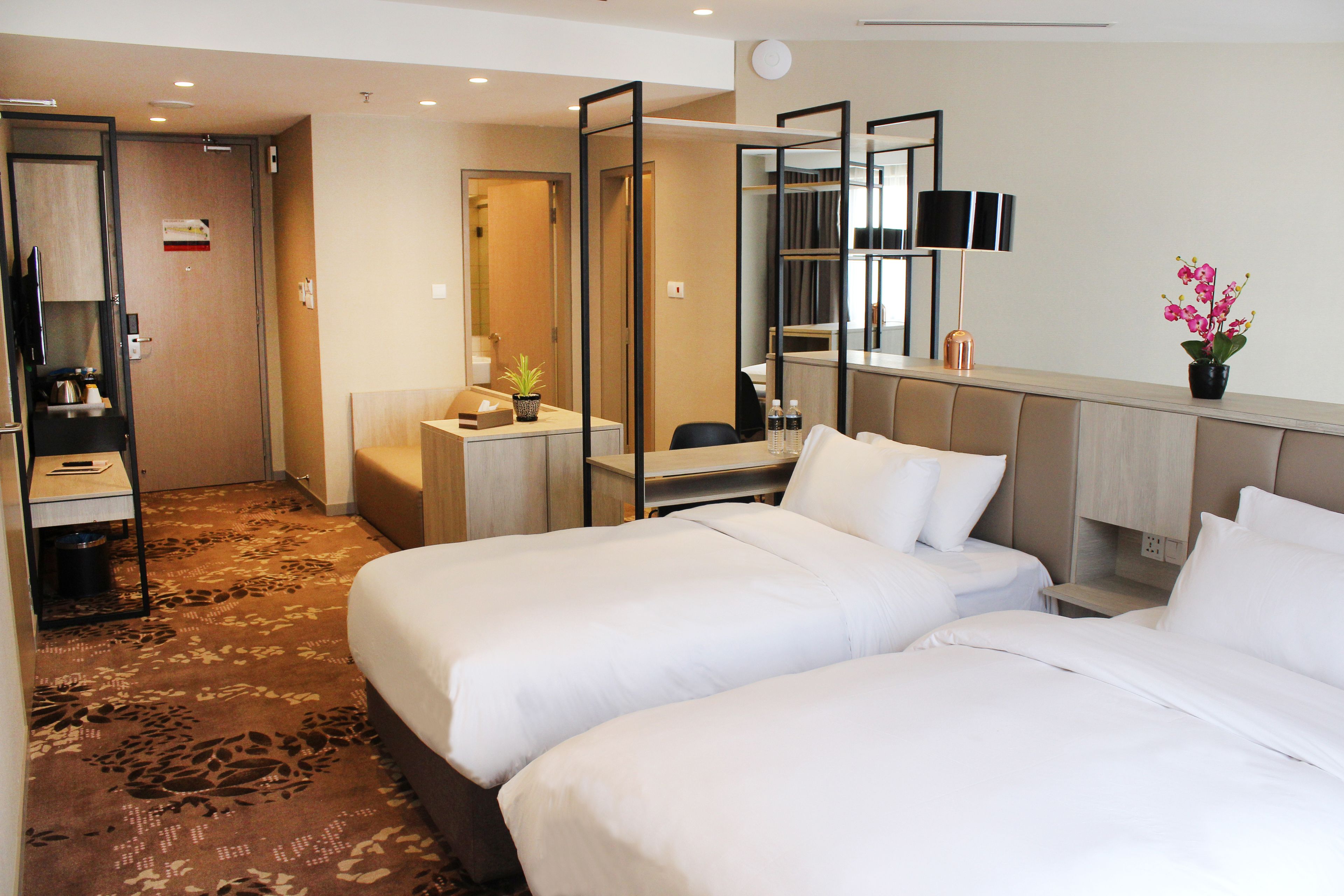 A hotel room with two beds, a desk, a chair, a mirror, a potted plant, and a small table with a lamp.