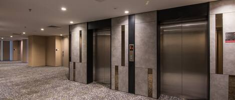 Un área de ascensores moderna con ascensores metálicos, una alfombra estampada e iluminación empotrada en el techo.