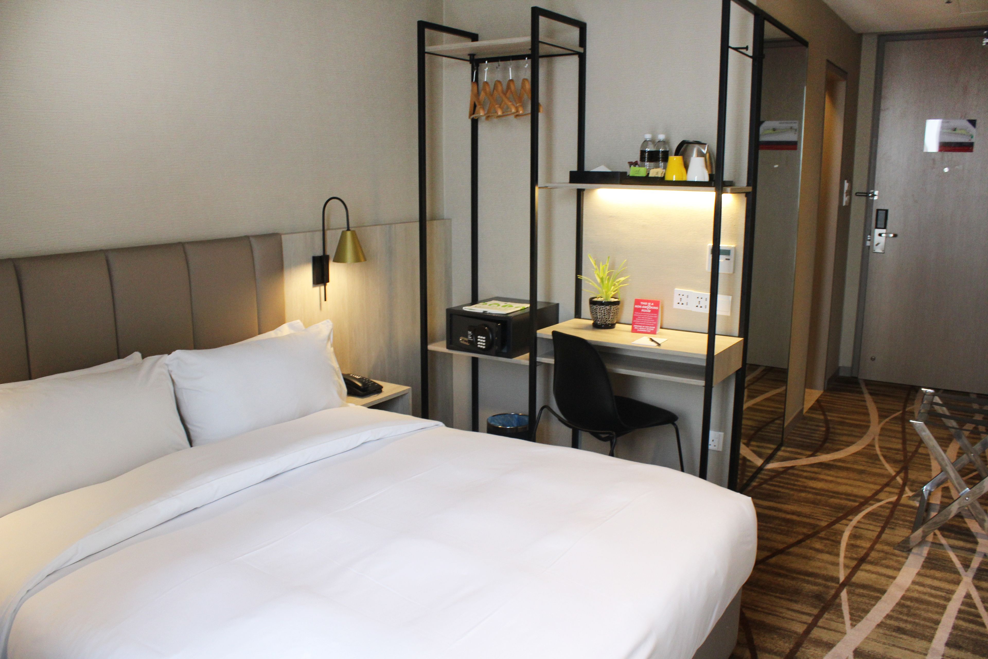 A hotel room with a bed, a desk with a chair, a shelf with toiletries, and a door.