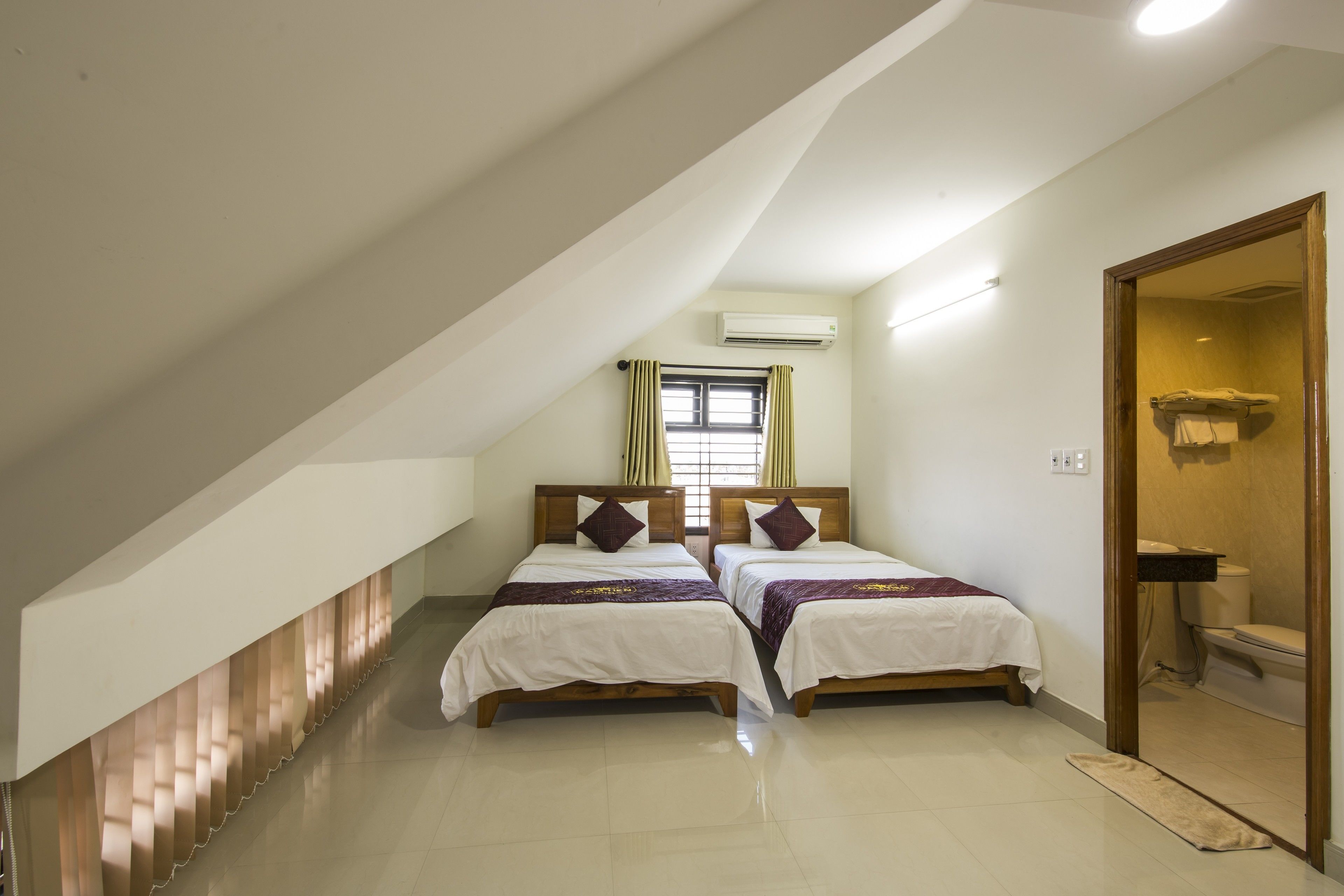 Un dormitorio con dos camas, una ventana con cortinas, un equipo de aire acondicionado y una puerta de madera que da a un baño.