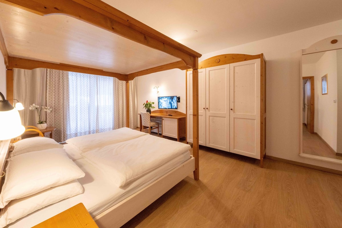 A hotel room with a large bed, a desk, a chair, a TV, and a wardrobe.
