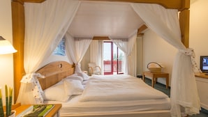 Ein Schlafzimmer mit einem hölzernen Kopfteil, einem Baldachinbett, einem Nachttisch mit einem Buch, einem Sessel und einem kleinen Tisch mit einem Korb.