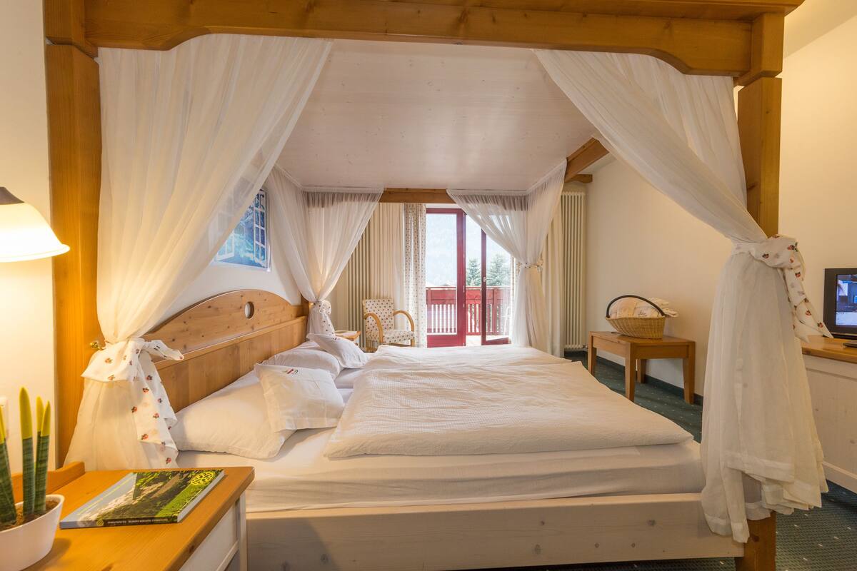 A bedroom with a wooden headboard, a canopy bed, a bedside table with a book, a chair, and a small table with a basket.