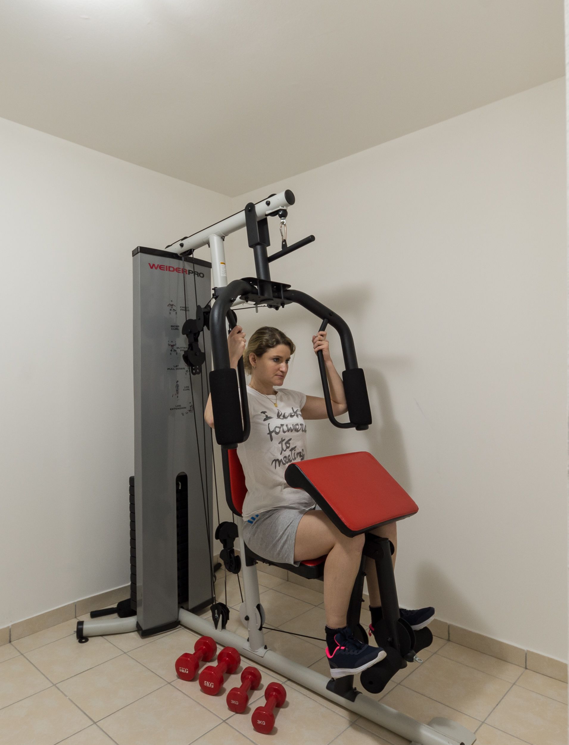 A person using a gym machine with a red seat and handles, surrounded by dumbbells on the floor.