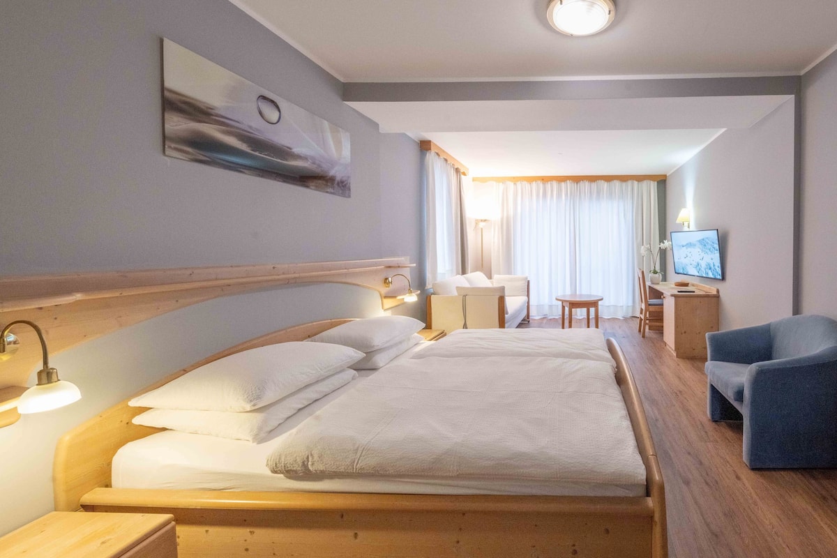 A hotel room with a bed, bedside table, lamp, chair, desk, TV, and curtains.