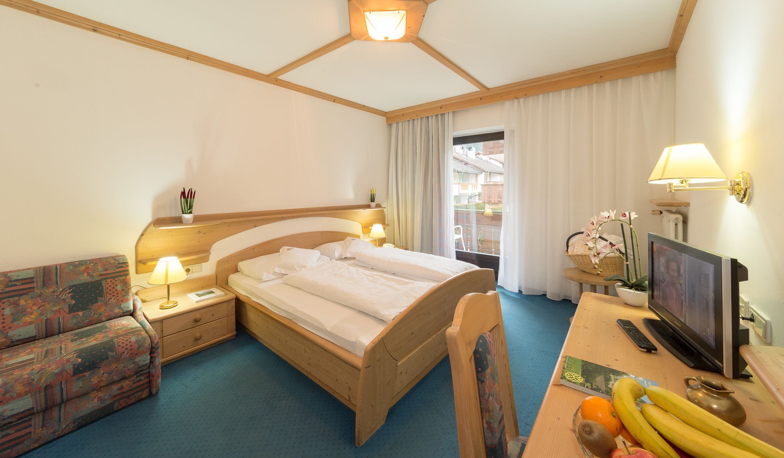Ein Hotelzimmer mit Bett, Schreibtisch inklusive Computer, Stuhl, kleinem Tisch mit Obst und Fernseher.