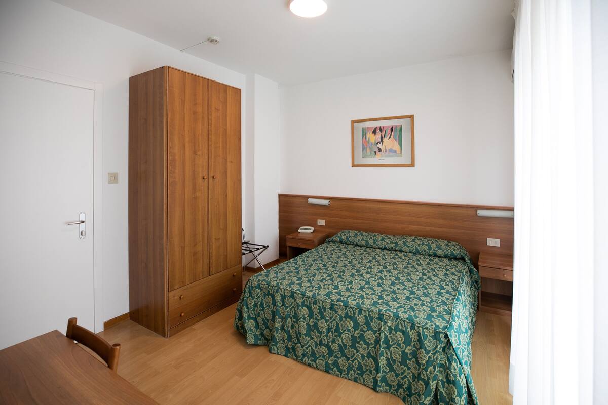 A bedroom with a wooden wardrobe, a bed with a patterned bedspread, a bedside table, and a wall-mounted artwork.