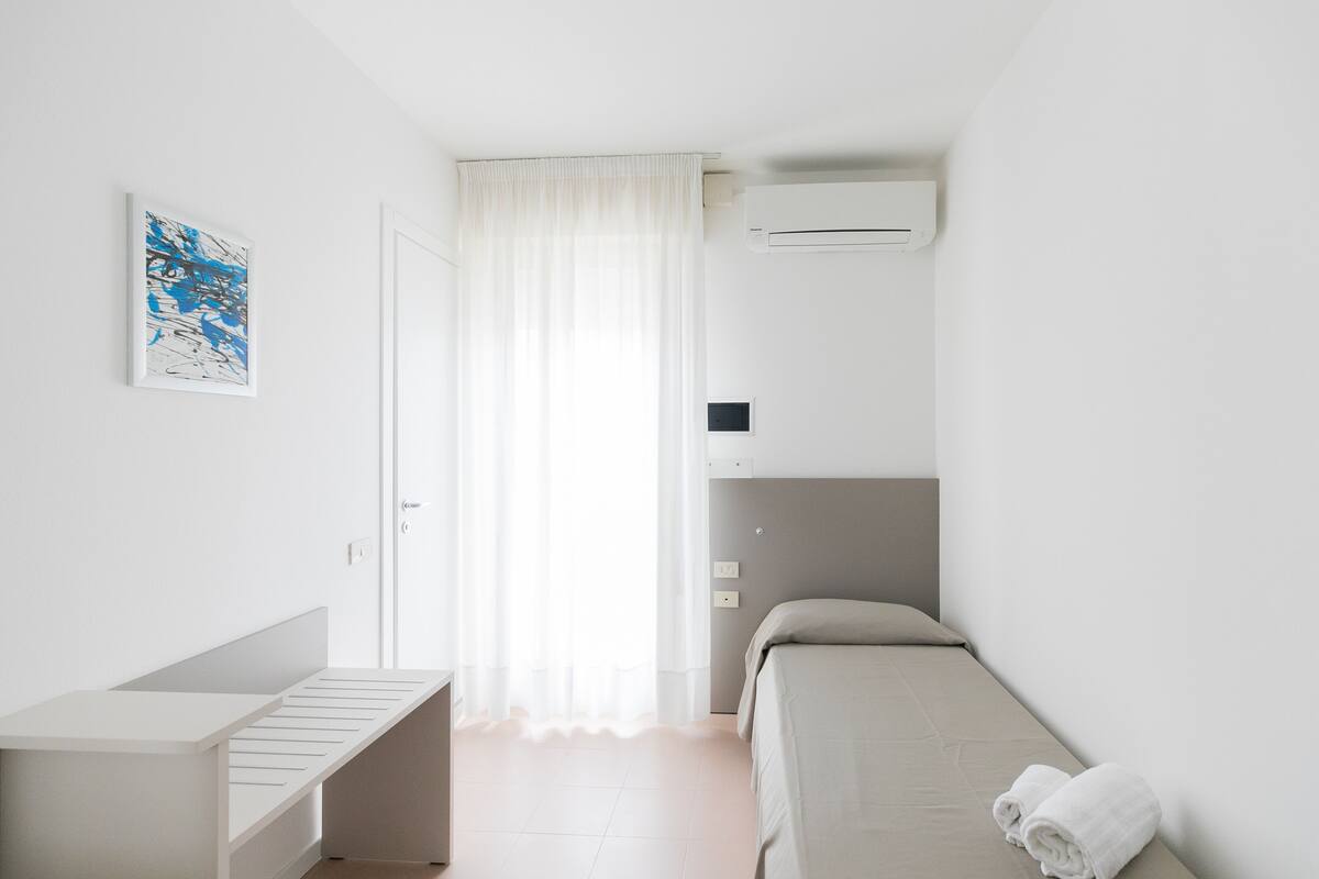 A small, modern bedroom with a bed, a desk, and a window with sheer curtains.