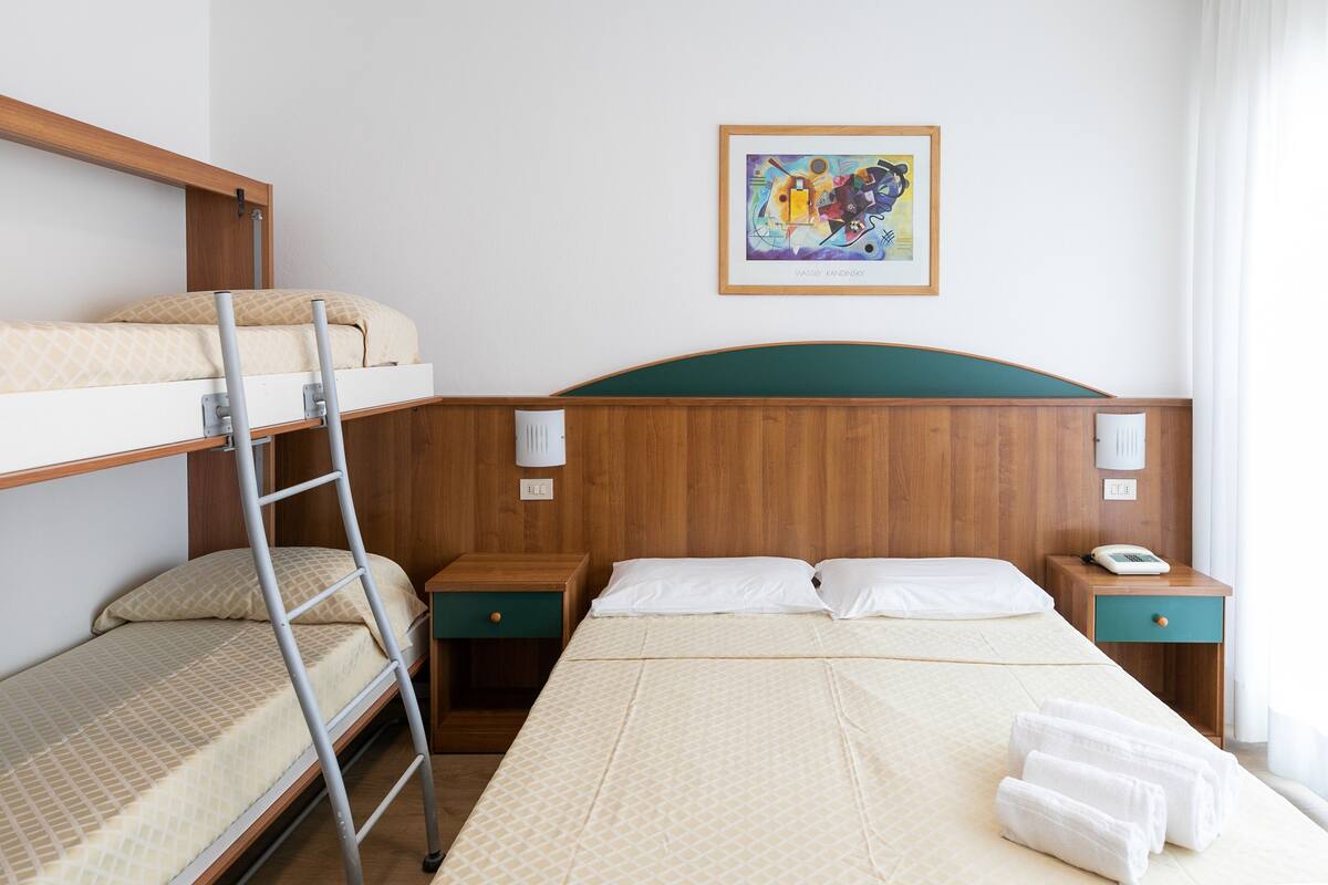 A hotel room with a bunk bed, a desk, a chair, a bedside table with a lamp, and a framed artwork on the wall.