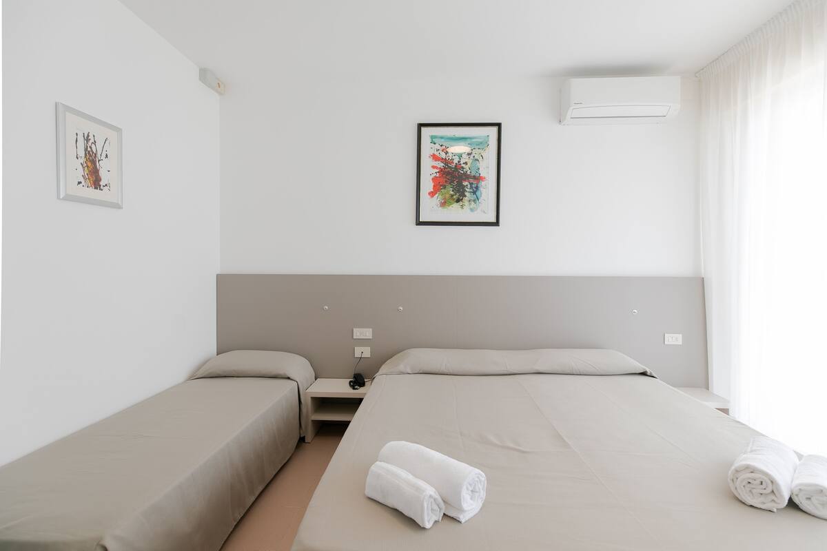 A hotel room with two single beds, a large bed, a nightstand, a framed picture, an air conditioner, and a window with curtains.