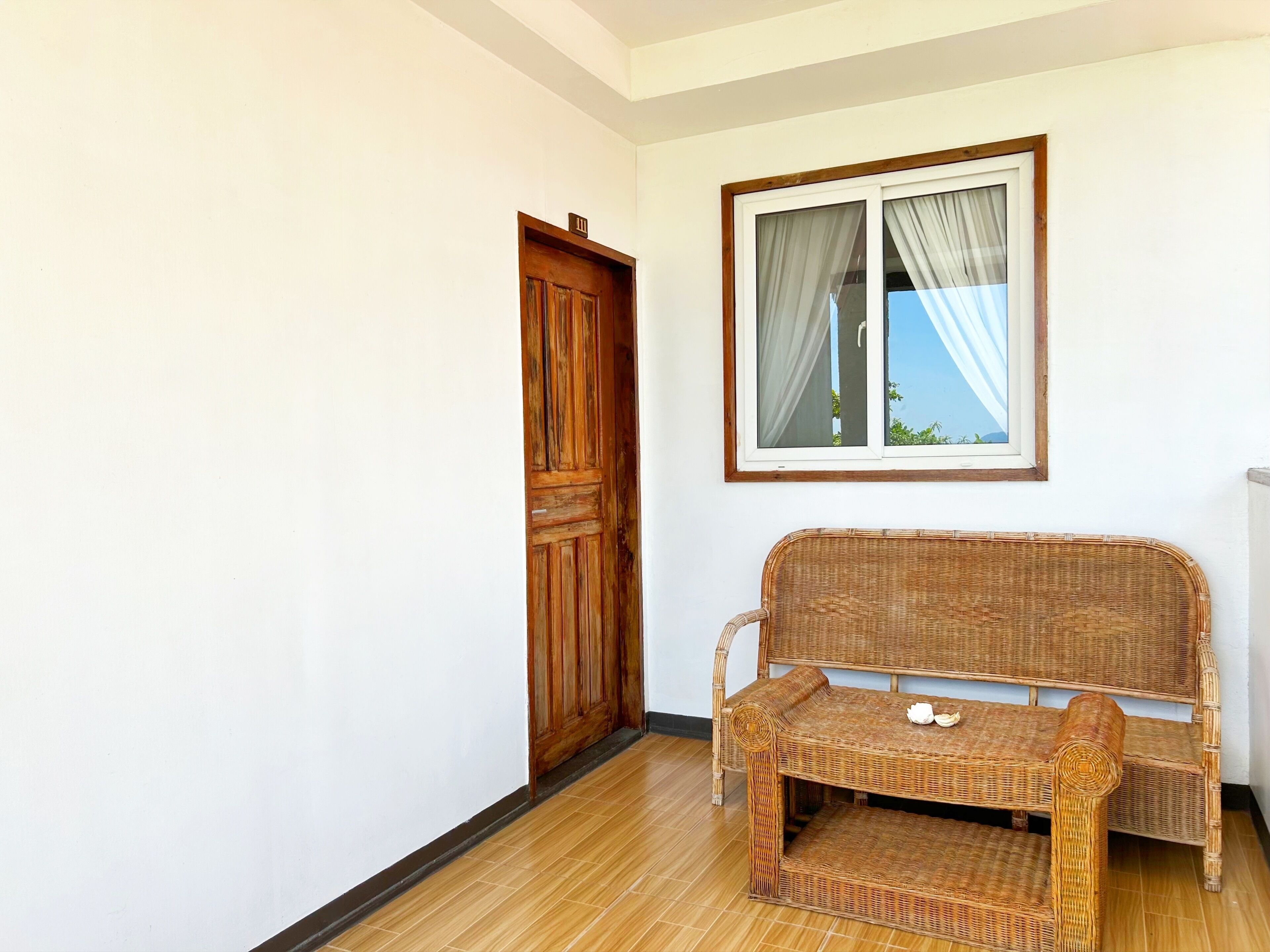 Una habitación con puerta de madera, un sofá de mimbre, una mesita con dos tazas y una ventana con cortinas.