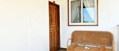 Una habitación con puerta de madera, un sofá de mimbre, una mesita con dos tazas y una ventana con cortinas.