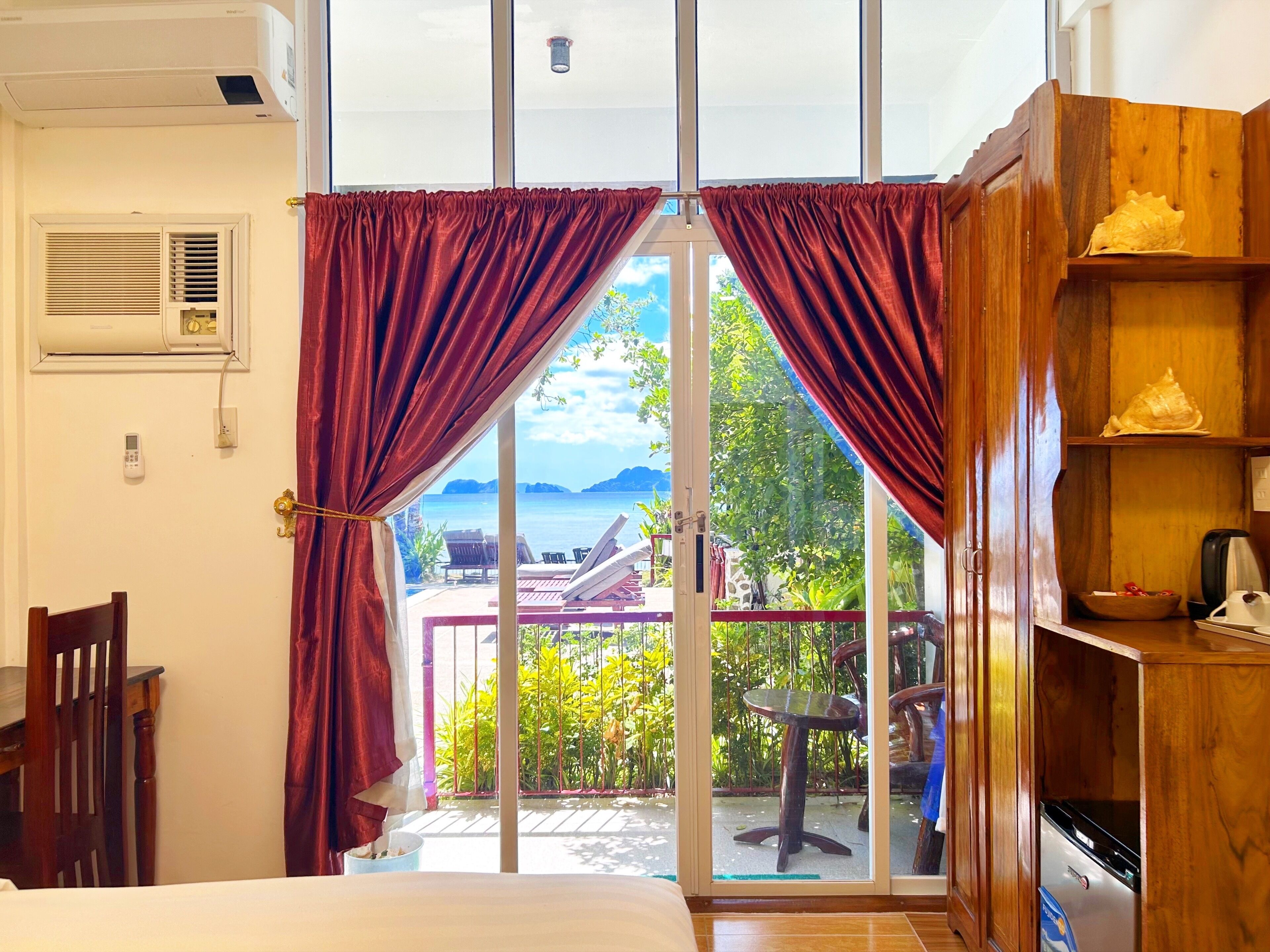 A room with a bed, a wooden chair, a table, a coffee maker, and a view of the sea through a glass door.