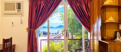 Quarto com cama, cadeira de madeira, mesa, cafeteira e vista para o mar através de uma porta de vidro.